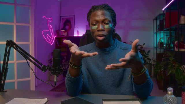 Medium UGC shot of cheerful black male influencer testing moisturizing eye patches on social media broadcast, while speaking to viewers about face massage and beauty procedures, looking at camera