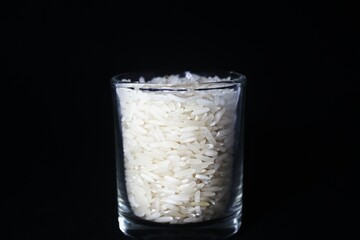 A clear shot glass filled with raw white rice grains. The shot glass is placed in the center against a solid black background, creating a strong contrast that emphasizes the texture and color of the r
