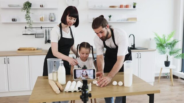 Caucasian family of bloggers filming cooking tutorial in kitchen. Parents teaching daughter baking. Whisking, kneading ingredients for dough in inviting atmosphere.
