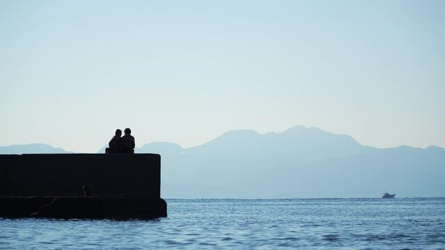 Loving couple sitting on a pier by the sea with a beautiful mountain landscape in the background.