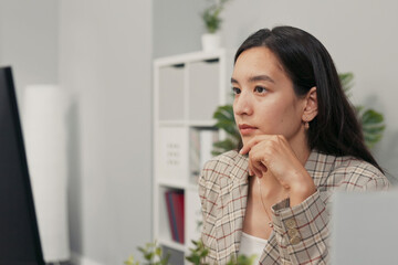 A focused pretty woman with long brown hair wearing a checkered jacket sits at desk in office in...