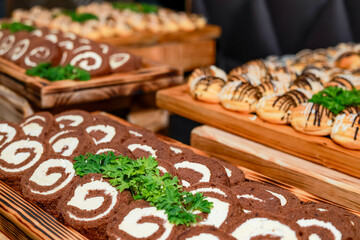 A wooden tray displays sweet treats, including chocolate rolls with white cream filling and cream puffs topped with chocolate glaze. Fresh cilantro garnish adds an appealing touch to the arrangement