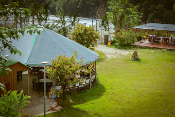 A circular structure covered with a dark green tarp serves as a seating area surrounded by greenery, with tables, chairs, and adjacent greenhouses visible in the serene outdoor setting