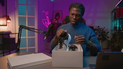 Medium UGC shot of cheerful black male influencer opening box with sports sneakers during live stream on social media, checking sole, admiring trendy design features, while looking at camera