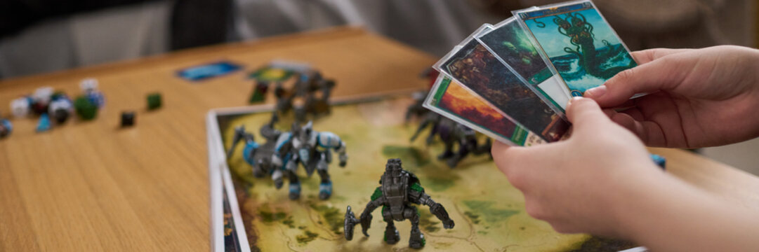 Header image of teenager holding game cards in hand while playing board game with friends at home copy space