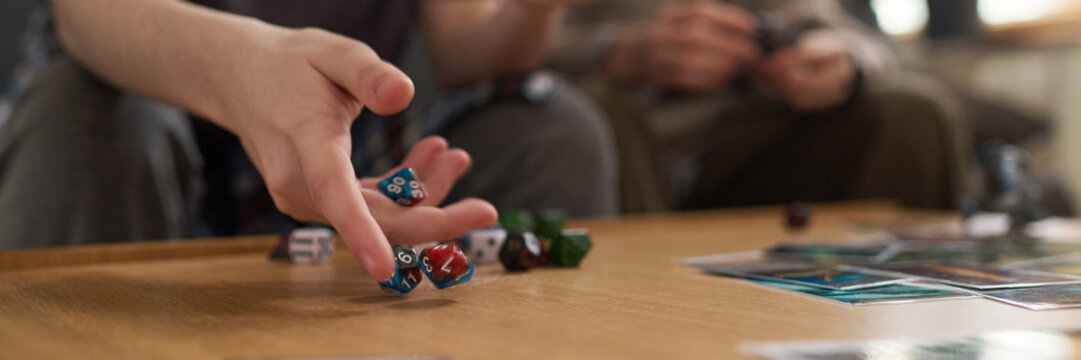 Header image of teenager throwing multi-sided dice on table while playing fantasy board game with friends at home or in dorm room copy space