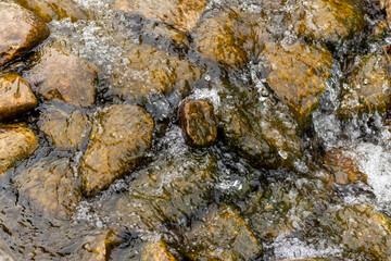 Clear water flows over weathered, brown rocks, creating dynamic ripples and white foam. The rocks vary in size and shape, displaying smooth, rounded surfaces, likely from erosion. 
