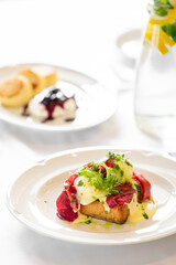 A close-up of a gourmet breakfast featuring toast with poached eggs, cured meat, greens, and hollandaise sauce, complemented by scones and a refreshing glass of water.