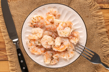 Seafood salad, shrimp with garlic on a ceramic plate with a knife and fork, with a jute napkin on a wooden table, close-up, top view.