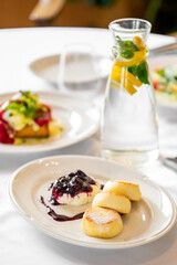Close-up of golden pancakes topped with berry sauce and cream on a white plate, surrounded by a refreshing carafe with lemon and mint, creating a gourmet dining ambiance.