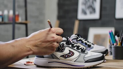 Close-up of hands painting on customized sneakers, the brush carefully outlines floral ornaments on the sneaker in a modern creative workshop