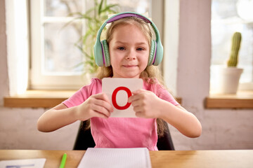 Little smiling pupil, girl presenting flashcard with red letter O while attending engaging online lesson in home setting. Concept of childhood, online education, remote studying
