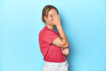 Blonde young woman in red t-shirt on blue background blink at the camera through fingers, embarrassed covering face.