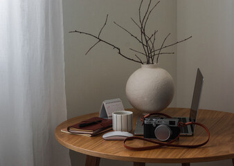 A laptop, a camera, a notebook, a cup of coffee, a ceramic round vase with branches on a round wooden table in the living room
