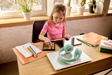 Girl using wooden abacus while studying at desk with digital tablet and school supplies in warm daylight interior. Math. Concept of childhood, online education, remote studying