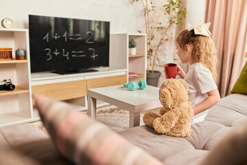 Girl watching math equations on TV with teddy bear and headphones on table, enjoying interactive...