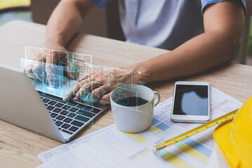 Person using laptop to design architectural floor plans digitally, with coffee, smartphone, and construction documents on desk.