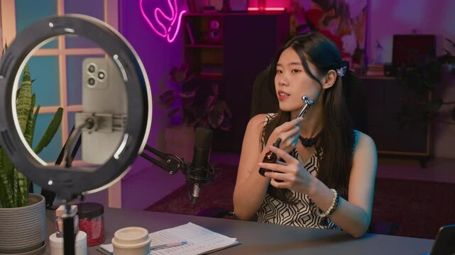 Medium shot of young Asian female beauty influencer sitting in front of microphone, ring light and smartphone in studio, filming face massage tutorial with roller and skin serum for social media