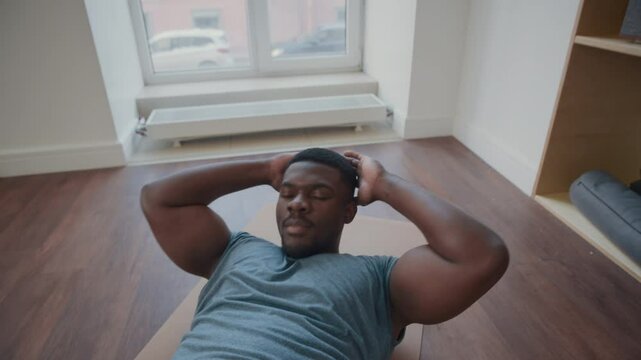 Muscular African American man in sports clothes doing curl ups with hands behind his head while exercising on mat in home gym