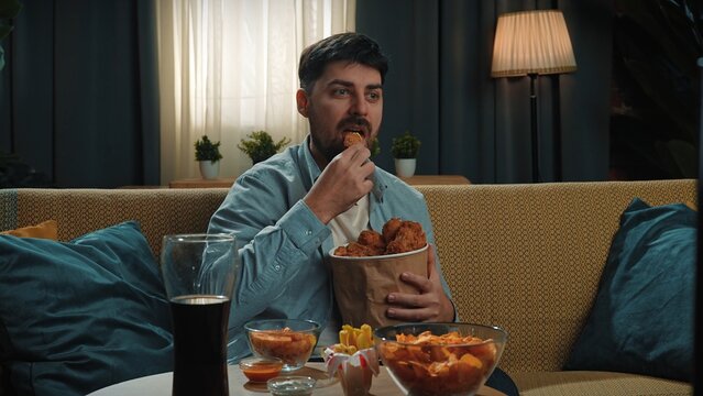 Man spending time at home with fast food delivery, sits on couch watching tv, dipping fried chicken leg in sauce and biting it, enjoying the taste.