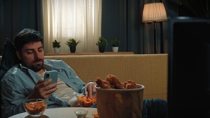 Bearded man spending time at home with fast food delivery, lays on couch, watching tv eating potato chips and scrolling smartphone.