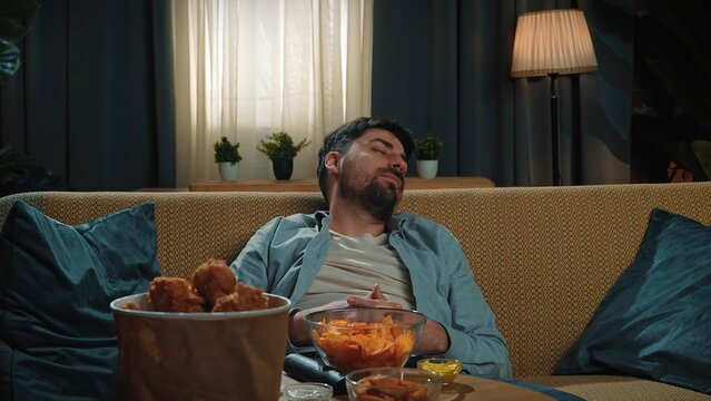 Bearded man spending time at home with fast food delivery, sits on couch and watching tv, getting sleepy yawning and falling asleep.