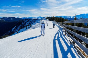 Obraz premium A skier in a white jacket and black pants is carving a turn on a groomed slope with a view of the snow-capped Alps in the distance. The sky is blue, and the sun is shining.