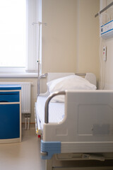 Empty hospital room with clean adjustable bed and blue bedside cabinet near window, white linens neatly arranged, bright sterile environment, elderly care, seasonal flu, patient recovery