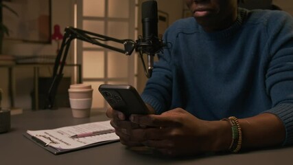Medium shot of midsection and lower face of anonymous black male blogger sitting at desk in studio in front of microphone, smiling while reading messages on smartphone before livestream