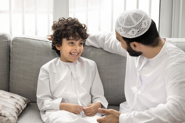 Joyful interaction between young Arabic father and his son, little cute boy dressed in traditional Muslim thobe, laughing enjoy conversation with dad seated on sofa at home. Family ties, strong bonds