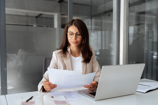 Concentrated latin business woman saleswoman working on laptop computer reading financial bank tax document in office workplace. Young manager businesswoman specialist doing bill paperwork using pc