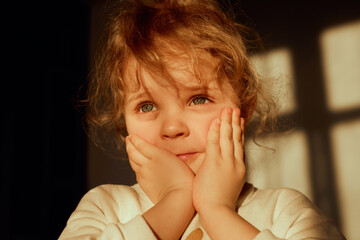 A young child with curly hair displays a pensive expression while resting hands on cheeks. The warm...