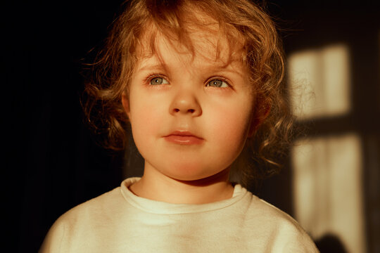A young child with curly hair and bright blue eyes stands in a softly lit room, gazing thoughtfully into the distance as warm golden light filters through the windows
