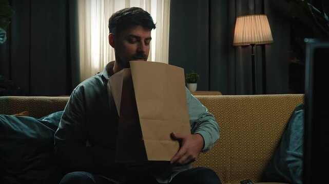 Adult bearded man spending time at home, sits on couch holding paper bag with fast food delivery, turns on tv and switches channels with remote. Slow motion.