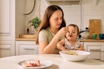 A mother gently feeds her baby in a sunny kitchen setting. The child eagerly opens their mouth for...