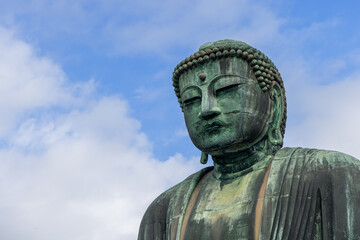 Serene face of the Kamakura Buddha framed by soft clouds with weathered bronze textures and sculpted details standing out against the vast sky