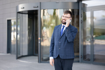 Fototapeta premium well-dressed businessman stands outside sleek office building smiling as he talks on his smartphone. contemporary architecture features revolving door and glass facade reflecting bright day.