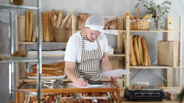 Man owner is busy checking paper documents, calculating income and expenses, expected increase in profit. Checks bill of lading. Bakery employee in apron examines documents, ponders debt restructuring