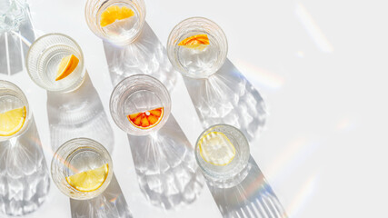 Fruit Infused Water in transparent Glasses with Shadows at sunlight on White background. Aesthetic view summer healthy drink with slices of lemon, orange with rainbow sun glare from glass cups, above