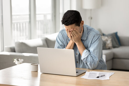 Man covers his face with hands while sitting at desk in front of laptop, got awful news, experiencing problems with device or internet connection, looking overworked, feels hopeless, tight deadlines