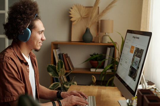 Side view of joyful young biracial man in headphones generating images using artificial intelligence on desktop computer