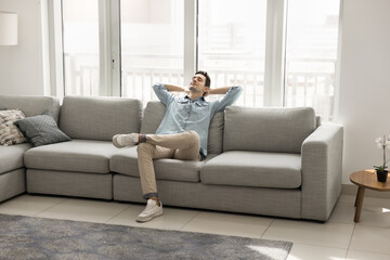 Peaceful young Hispanic man dressed casually reclining on cozy sofa in contemporary living room, enjoying moment of tranquility and calmness on carefree weekend leisure at home. Relaxation and comfort