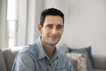 Portrait of smiling Hispanic man dressed in casual shirt posing for camera sit on couch at home,...