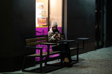 Lonely night in the city as a young woman sits on a bench engrossed in her phone illuminated by a colorful window glow while waiting for a friend