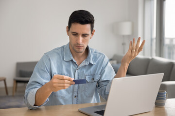 Hispanic man sit at table with laptop, holding card with concerned expression, experiences payment issue, transaction declined, feeling worried about insufficient funds, blocked card, or banking error