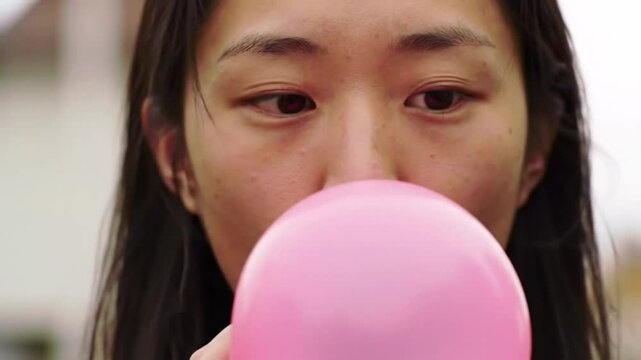 A young asian woman with dark hair blowing a pink bubble, captured in a close-up shot, showing her face and the inflated bubblegum.