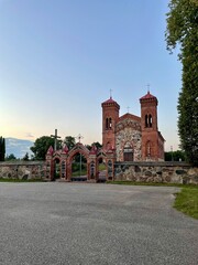 Obraz premium Karvys Church of St. Joseph, Lithuania. Red-brick church with twin towers and crosses, set behind a stone wall. Gothic architectural style with arched windows and entrance. 