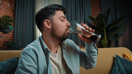 Bearded man spending time at home with fast food delivery, sits on couch and drinking cola from a large glass, taking a sip and enjoying the taste. Slow motion