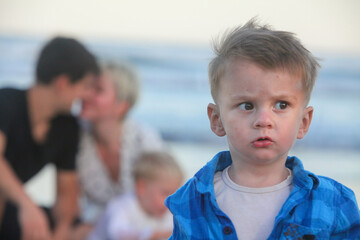 closeup portrait of anxious serious toddler with his cheerful family in background on the beach at...