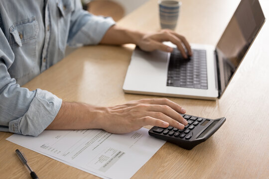 Cropped view man managing finances seated at desk, using laptop and calculator, researching, analyzing data, or accessing financial software or spreadsheets, budgeting, balancing incomes and expenses